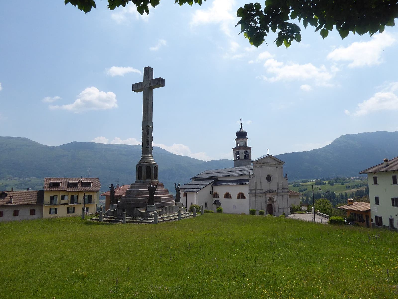 Santa Croce del Bleggio con la croce sul prato, la chiesa e le montagne