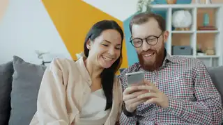 Coppia sorridente sul divano di casa mentre usa lo smartphone
